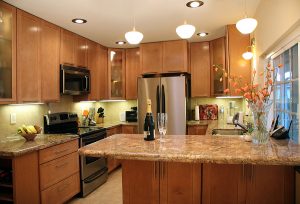 A modern kitchen features wood cabinets, granite countertops, pendant lighting, and stainless steel appliances. There is a bottle of champagne and two glasses on the counter.