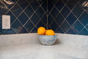 A close-up image of quartz countertops and a blue tile backsplash. A small bowl of oranges is in the middle of the image.