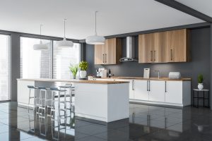 Corner of white wooden kitchen with panoramic view. White lower cabinets. Big table with wooden worktop. Blue bar stools and grey interior. Three lights above the table