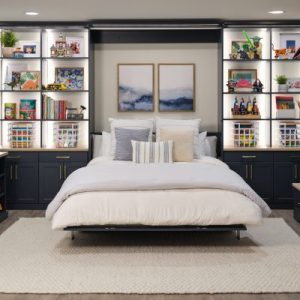 A room with lit shelving, cabinetry, and a made up murphy bed that has pillows and bedding